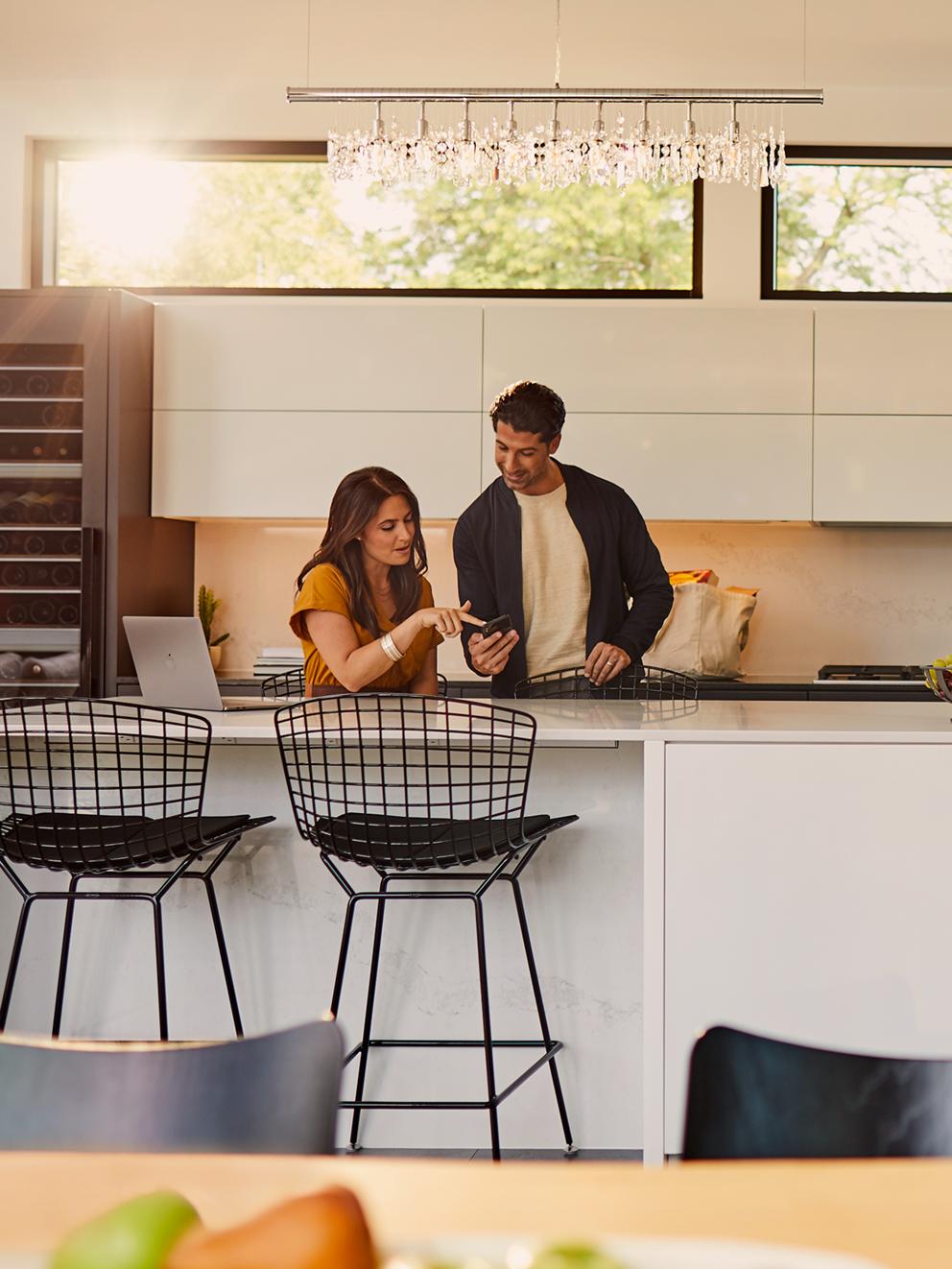 A man and woman standing in the kitchen and lookin at a phone.