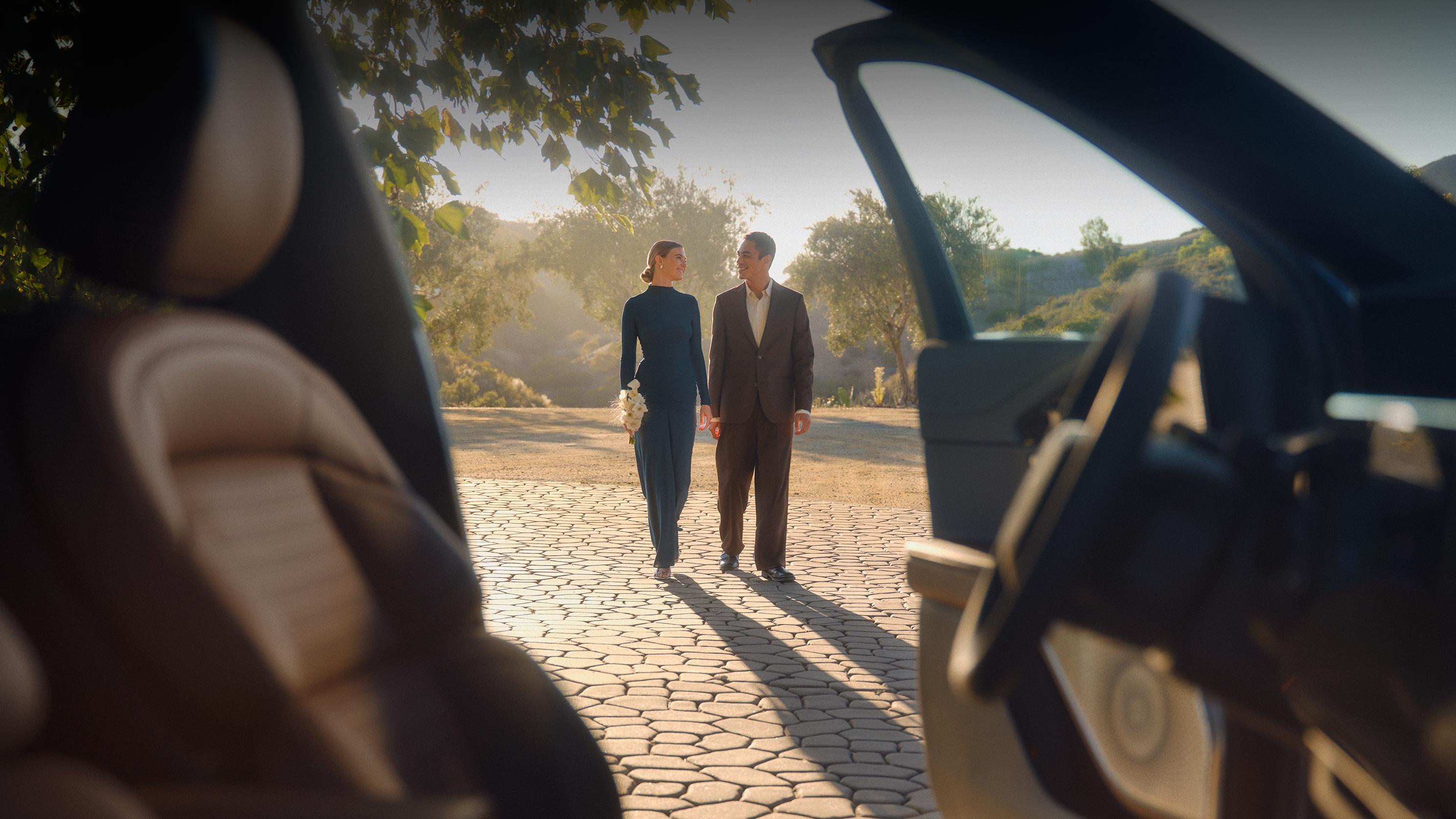 A man and a woman holding hands and smiling at each other while walking outdoors, viewed from inside a car.