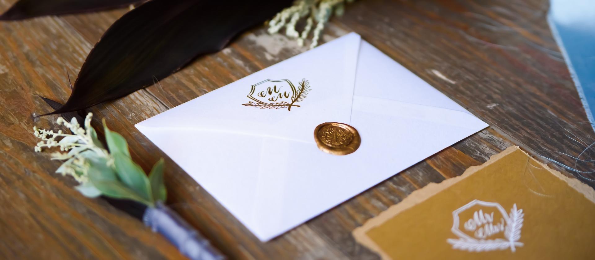 An envelope with a decorative seal holding an invitation lays on a wooden surface with floral arrangements